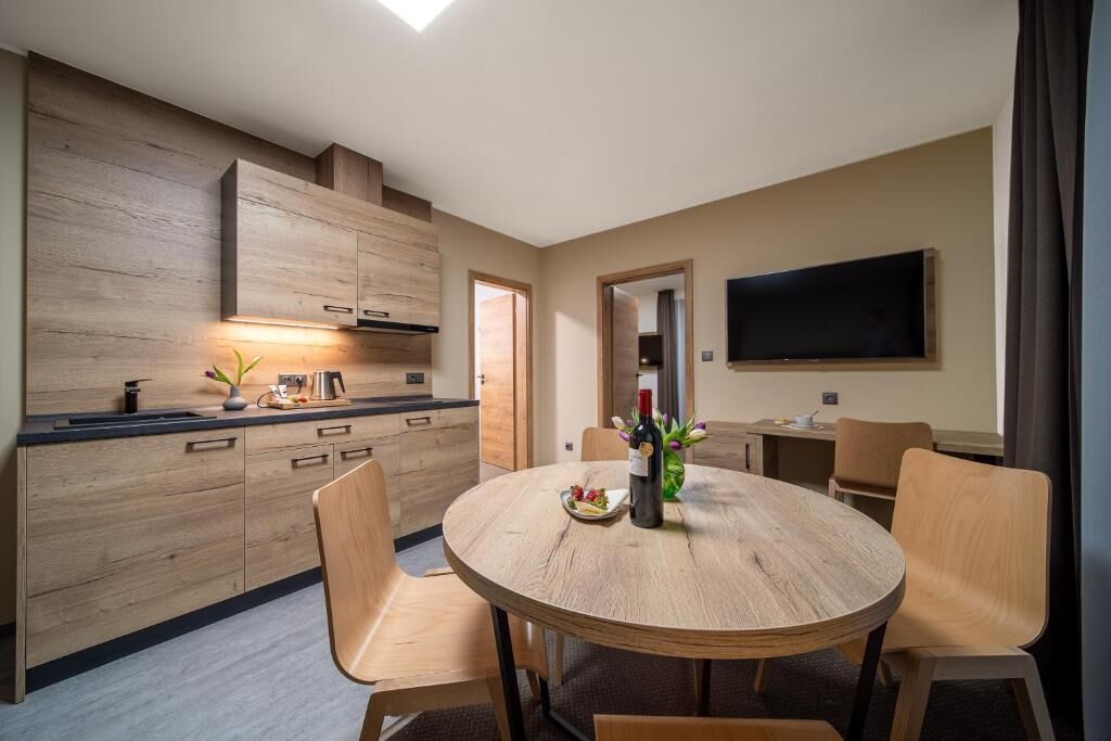 A modern kitchen with wooden cabinets, a dining table with chairs, and a flat-screen TV mounted on the wall.