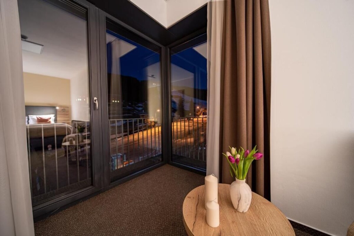 Et hotelværelse med en skydedør til en balkon, et rundt træbord med stearinlys og en blomstervase, samt udsigt over bybilledet om natten.