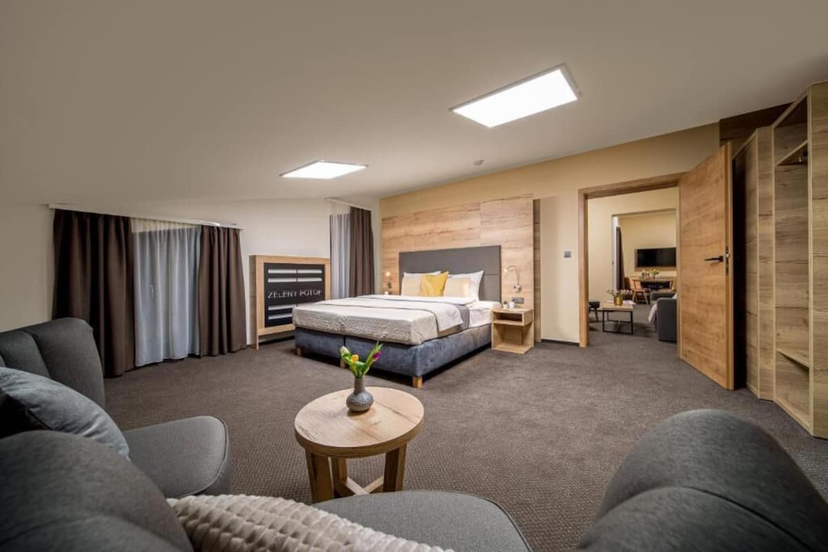 Una habitación de hotel moderna con una cama grande, una zona de estar con una mesita y un salón separado con televisor.