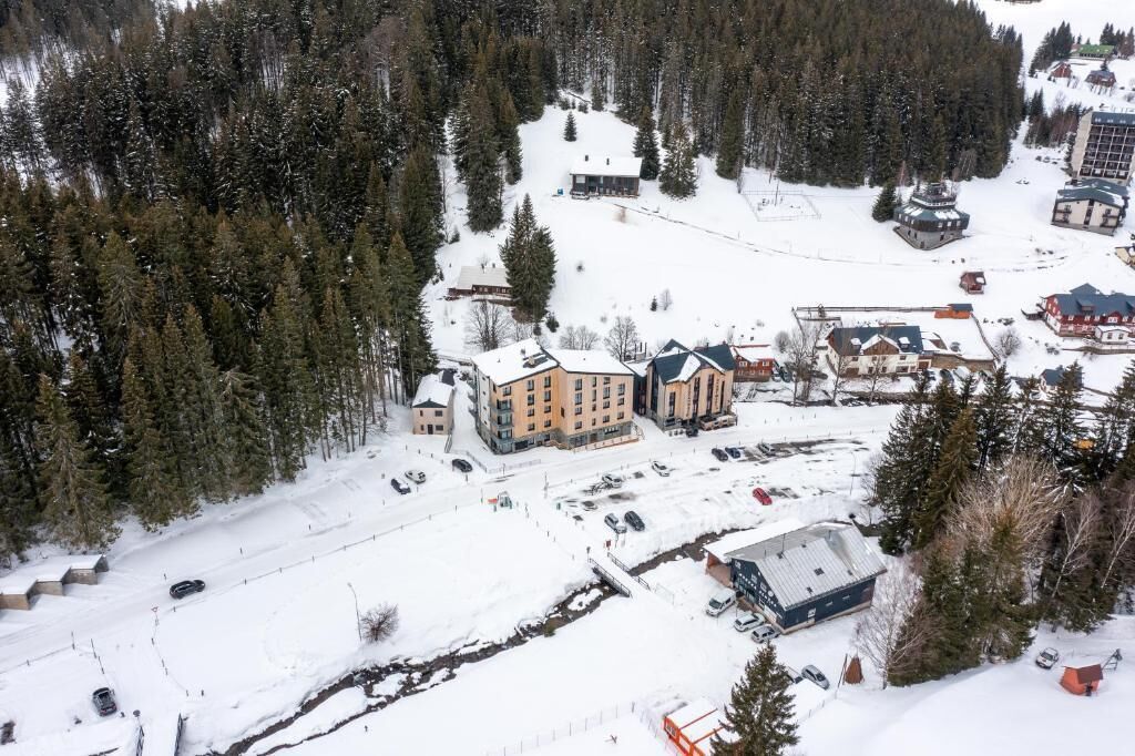 Vista aérea de un complejo de esquí con varios edificios, incluyendo un gran lodge y varias estructuras más pequeñas, rodeado de bosques densos y terreno cubierto de nieve.