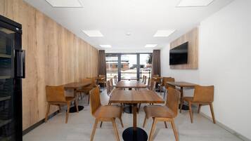 Un espace repas moderne avec des tables et des chaises en bois, un téléviseur à écran plat fixé au mur et une vue sur l’extérieur grâce à de grandes fenêtres.