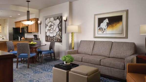 A hotel room with a sofa, a dining table with chairs, a kitchen area, and a large flower painting on the wall.