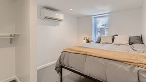 A bedroom with a bed, a window with blinds, an air conditioning unit, and a bedside table with a lamp. - Pet Friendly I Remodeled I Near RiNo | Sleeps 7 (Denver)