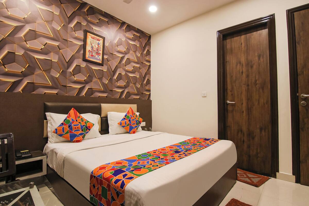 Quarto de hotel com design moderno, apresentando uma parede com padrão geométrico, uma cama grande com almofadas coloridas e uma porta de madeira.