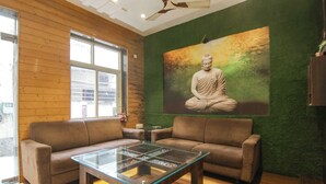 Une pièce avec deux canapés en cuir marron, une table basse en verre, un plancher en bois, un mur vert orné d’une statue de Bouddha et un ventilateur de plafond.