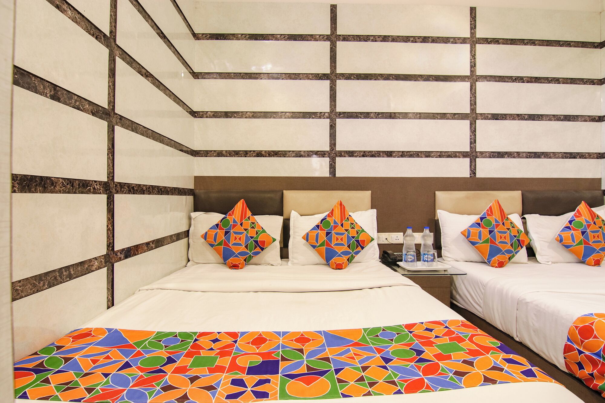 A hotel room with two beds, a large colorful geometric-patterned bedspread, and a headboard with a geometric design.