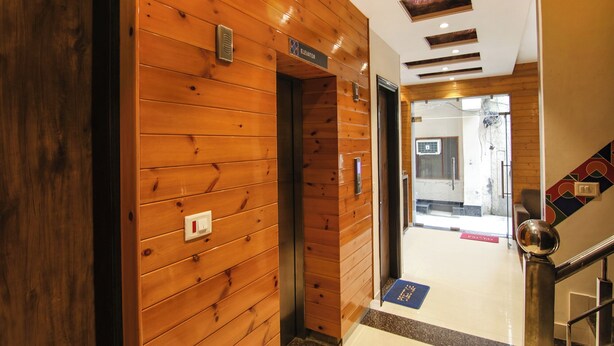Un couloir intérieur en bois, avec un panneau indiquant la présence d’un ascenseur, une porte en verre donnant sur une autre pièce et un escalier en métal muni d’une main courante.