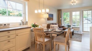 A modern kitchen with a dining area, wooden chairs, and a sofa. There is a window with blinds, a lamp, and a painting on the wall. - Strandnahe 3-zimmer Ferienwohnung auf ca. 55m² Wohnfläche, für bis zu 4 Personen (Wenningstedt-Braderup (Sylt))