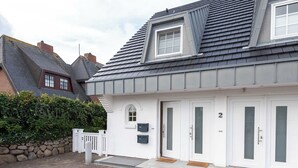 A two-story house with a grey roof, white exterior walls, and a small dormer window. There are two doors, each with a number above it, and a small porch area with steps. - Strandnahe 3-zimmer Ferienwohnung auf ca. 55m² Wohnfläche, für bis zu 4 Personen (Wenningstedt-Braderup (Sylt))