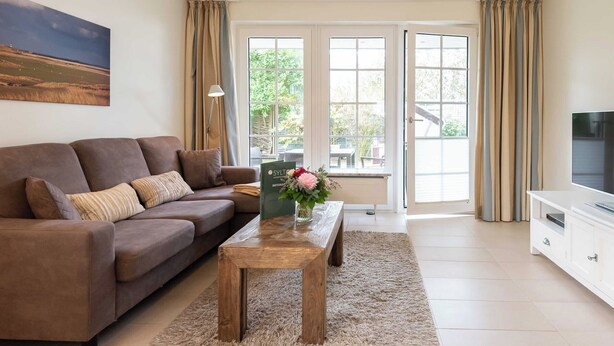 A living room with a brown sofa, a wooden coffee table, a TV stand, and a large window with curtains. - Strandnahe 3-zimmer Ferienwohnung auf ca. 55m² Wohnfläche, für bis zu 4 Personen (Wenningstedt-Braderup (Sylt))