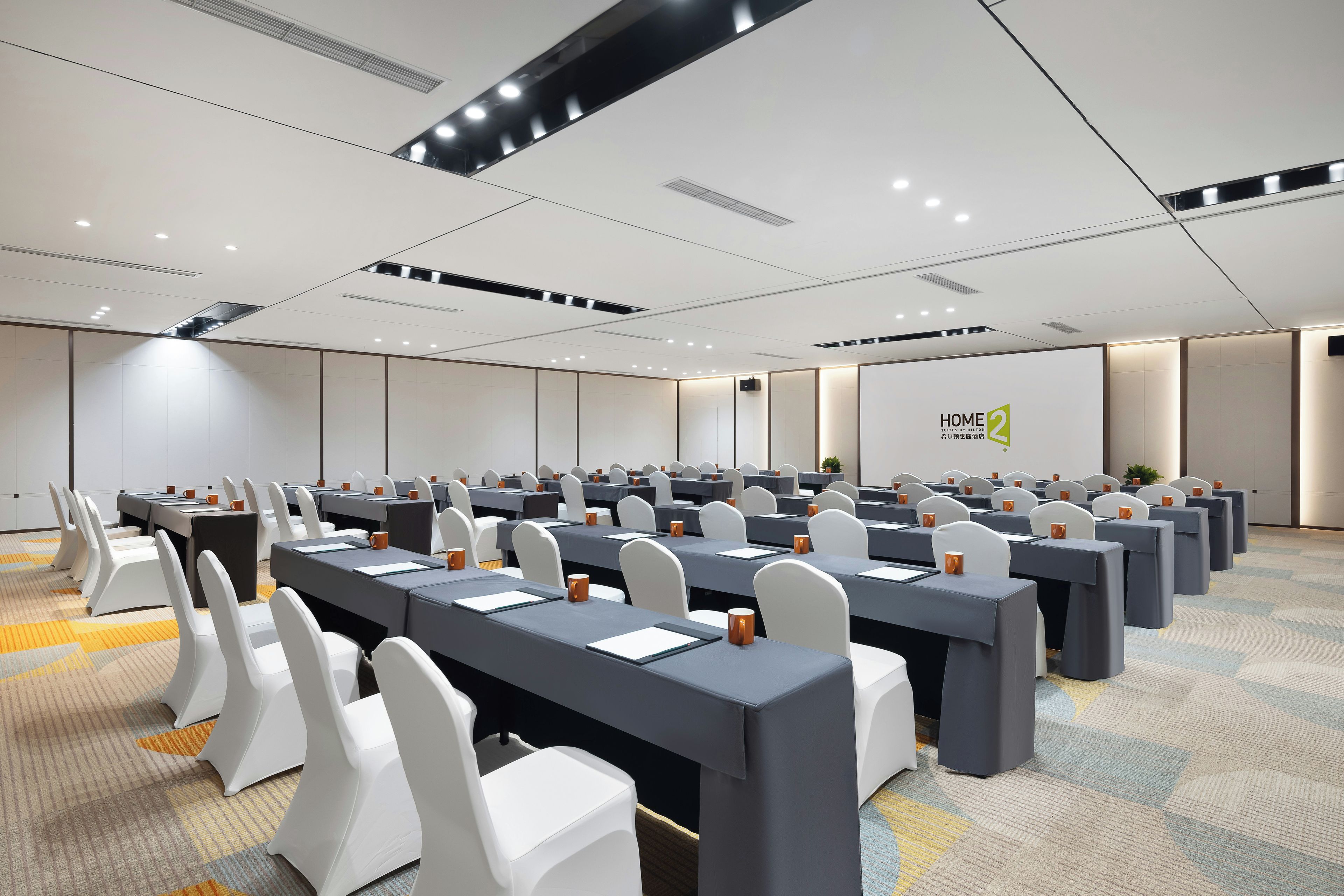 Une salle de conférence avec des rangées de tables et de chaises, un écran de projection et un panneau indiquant « HOME 2 Suites by Hilton ».