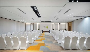Un salón de conferencias con filas de sillas blancas, piso alfombrado y una pantalla grande con el logotipo de HOME 2.