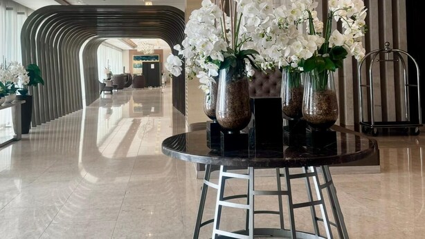 A modern lobby with a curved reception desk, a central round table with vases of white orchids, and hanging pendant lights. - MOA Staycation 10mins to Airport. 10mins walk to iconic Mall of Asia. (Pasay)