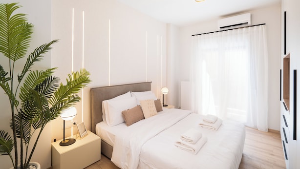 A modern bedroom with a large bed, bedside lamps, a potted plant, and a view of the outdoors through sheer curtains.