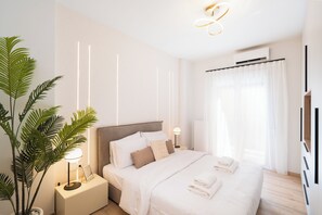 Quarto moderno com uma cama grande, luminárias de cabeceira, um vaso de plantas e vista para o exterior através de cortinas leves.