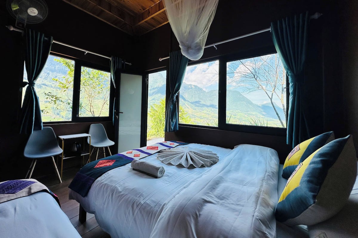 A bedroom with a bed, a desk, and two chairs. There are large windows with a view of mountains.
