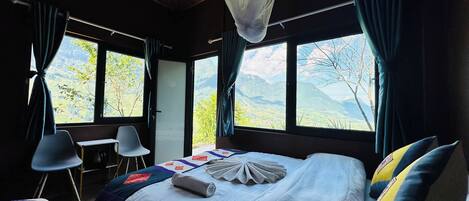 Un dormitorio con cama, un escritorio y dos sillas. Amplias ventanas ofrecen vistas a montañas.
