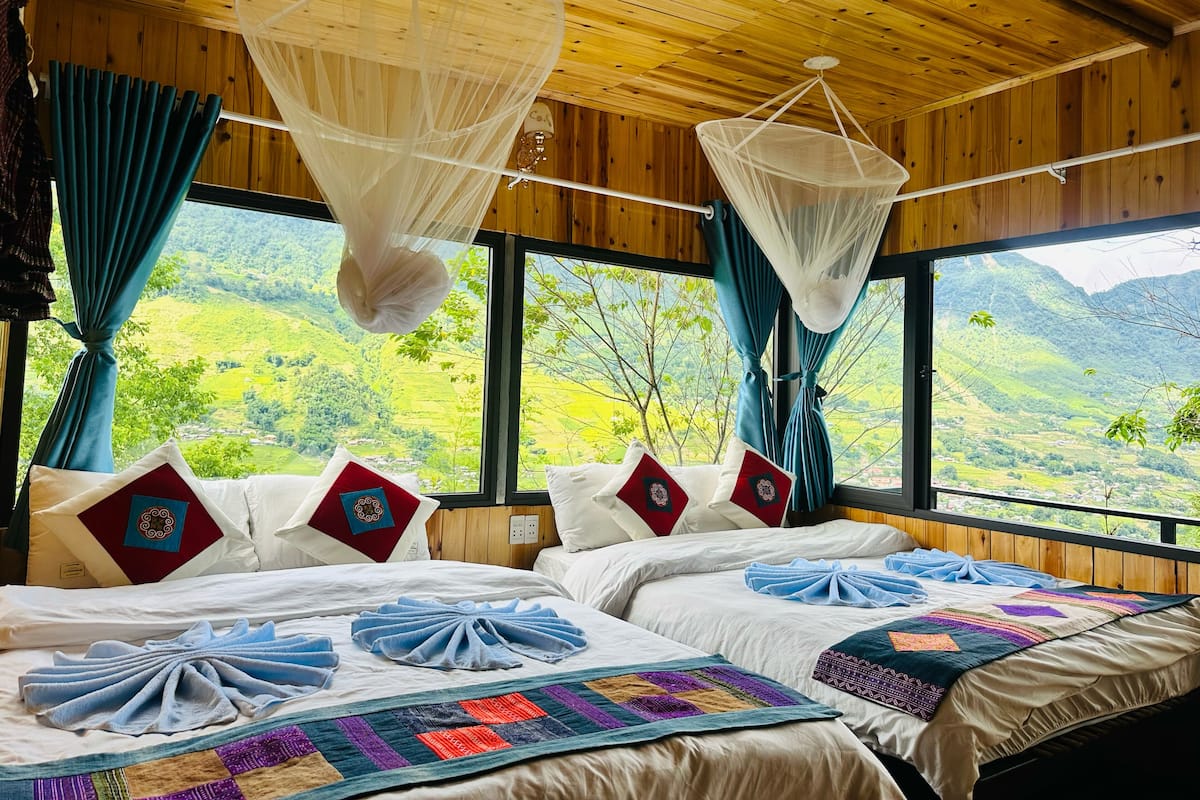 A room with two beds, large windows, and a view of green hills.