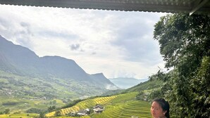 A person sitting on a chair with a table, overlooking a valley with terraced fields and mountains in the background. - Dinh Sapa Trekking & Homestay (Ta Van)