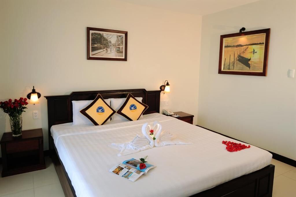 A neatly made bed with a swan and heart design, two pillows, a vase of red flowers, and a framed picture on the wall.