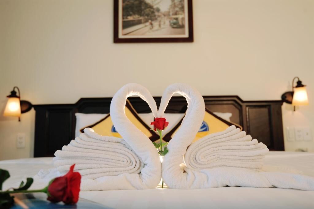 Una cama bien hecha con toallas dispuestas en forma de corazón, una rosa roja y un cuadro en la pared.