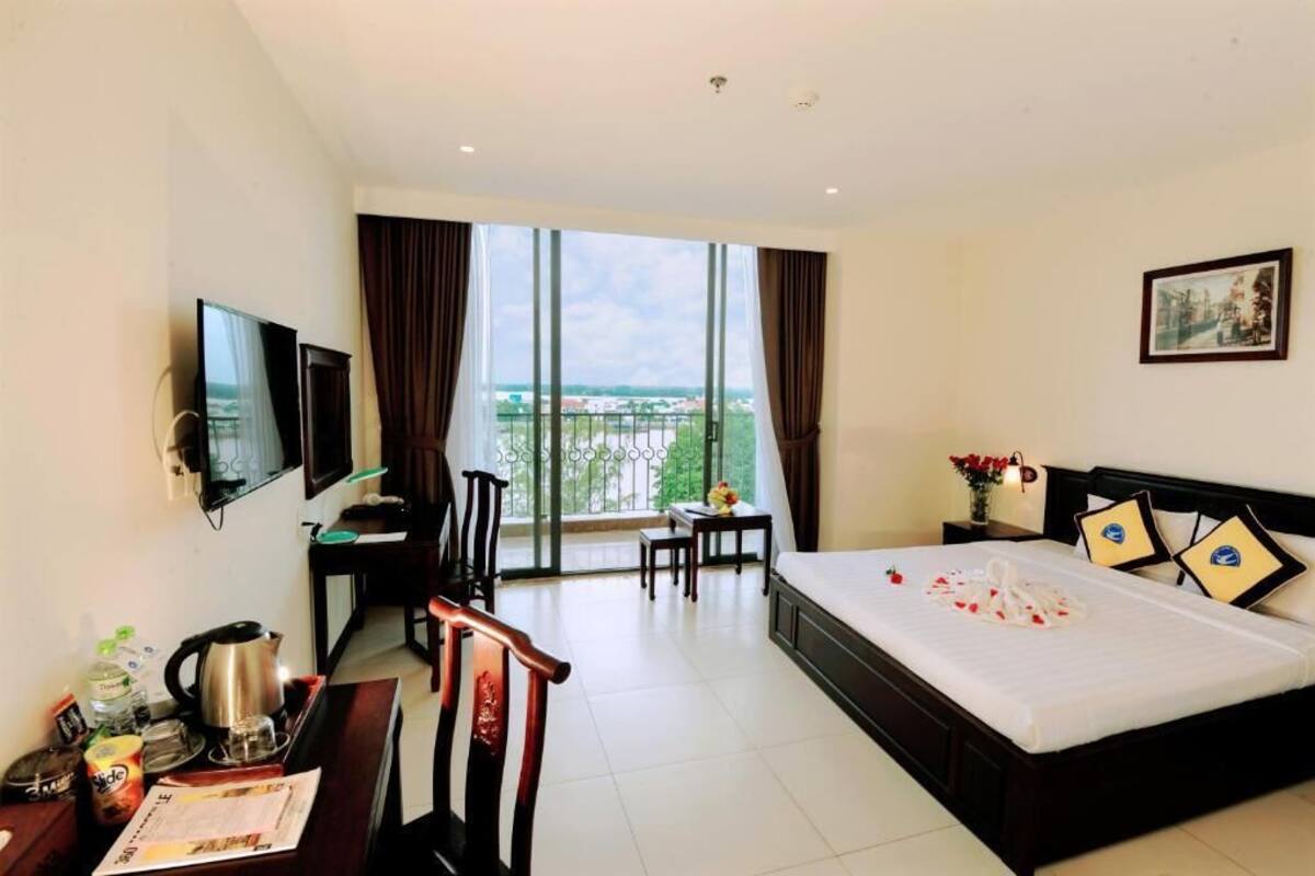 A hotel room with a large bed, a desk with a chair, a television, and a balcony with a view.