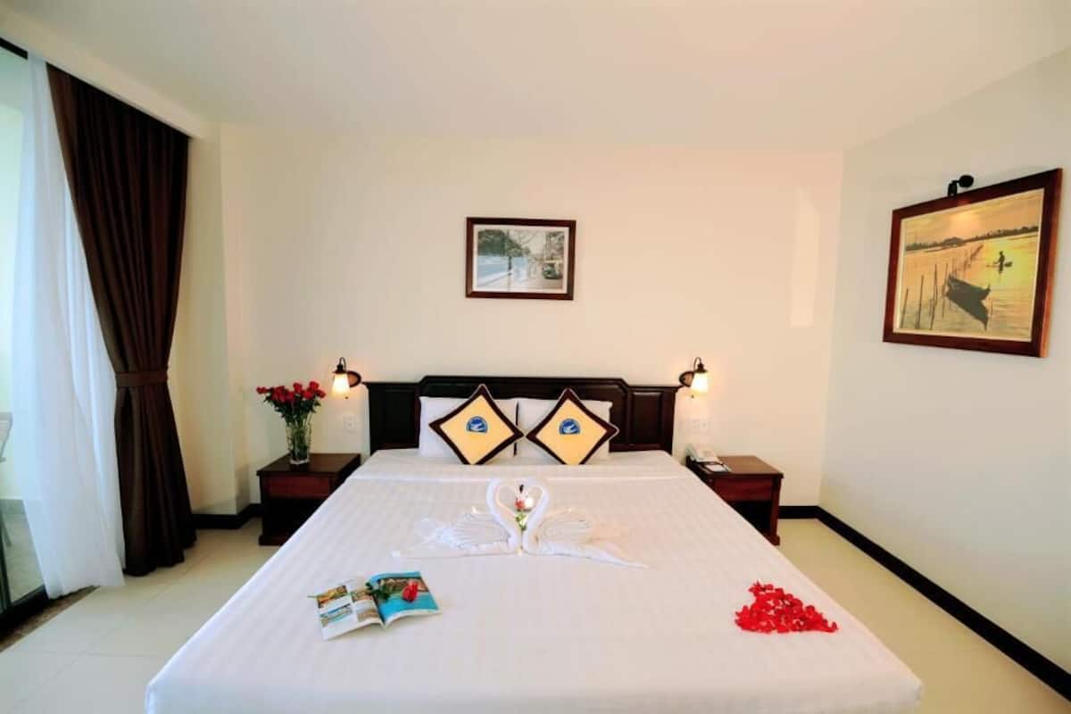 A hotel room with a neatly made bed, two pillows with a logo, a framed picture on the wall, and a nightstand with a lamp.