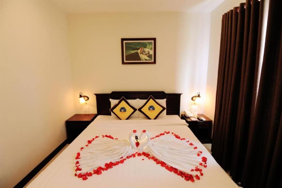 A hotel room with a bed, two nightstands, a headboard with decorative pillows, a framed picture on the wall, and a curtain.