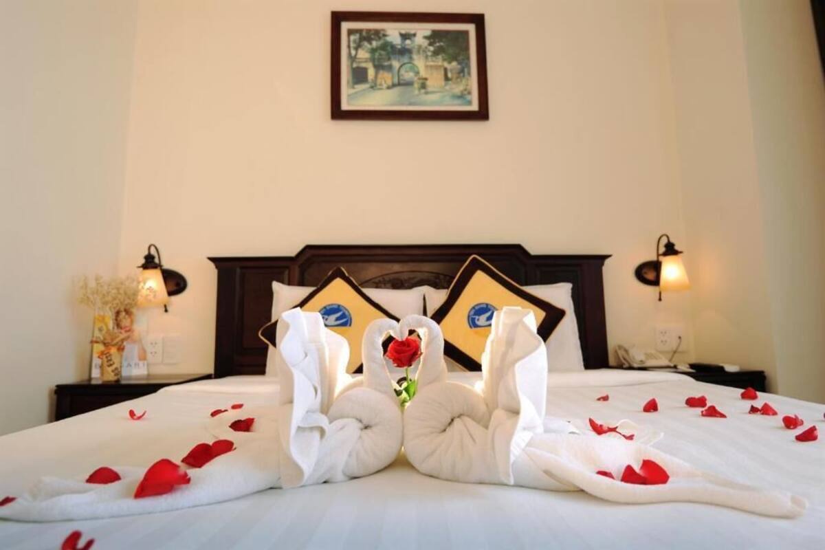 A neatly made bed with a rose and swan-shaped towels, a framed picture on the wall, and bedside lamps.