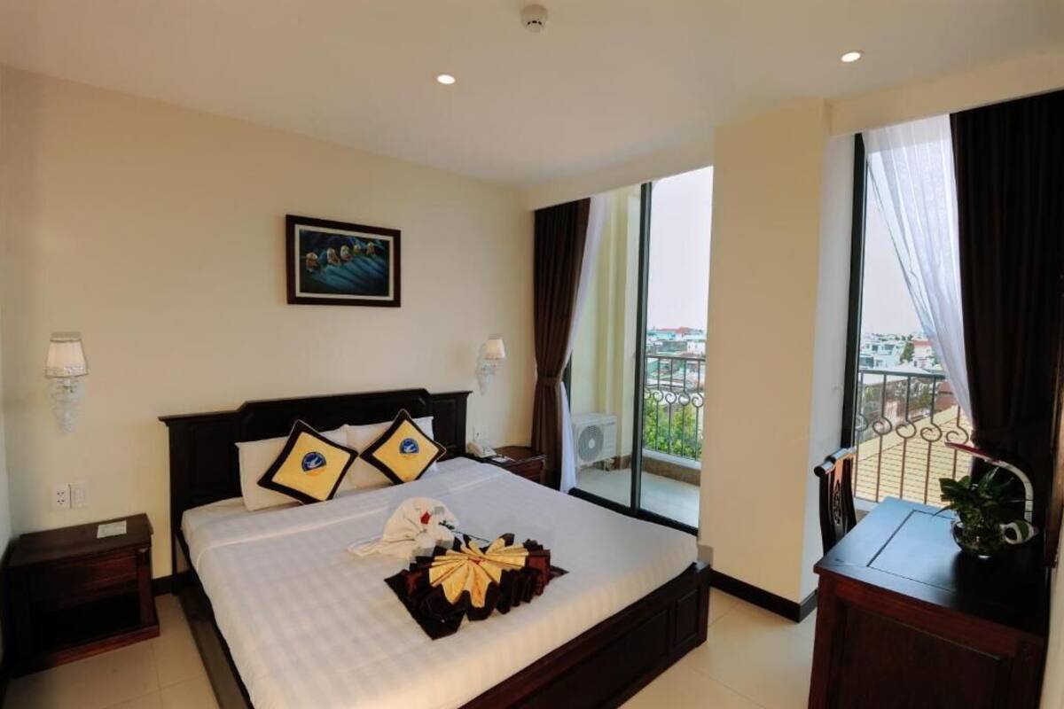 A hotel room with a large bed, a view of the city through the window, and a balcony with a railing.