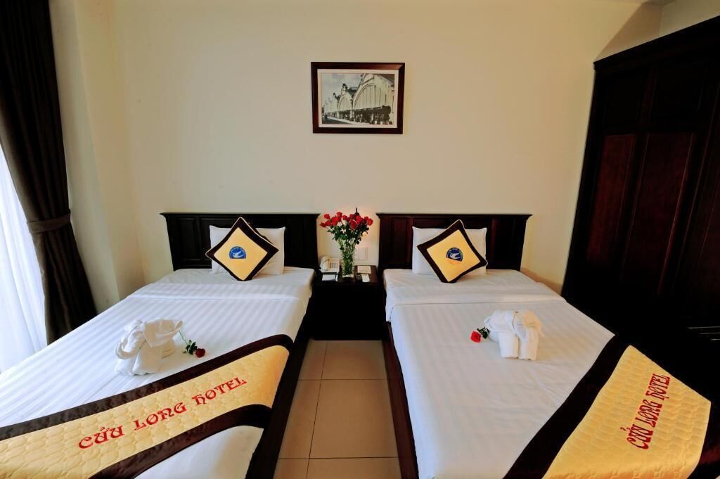 Une chambre d’hôtel avec deux lits, chacun étant agrémenté d’un oreiller et d’une petite table sur laquelle est posé un vase de fleurs. Un tableau encadré est accroché au mur, au-dessus des lits.
