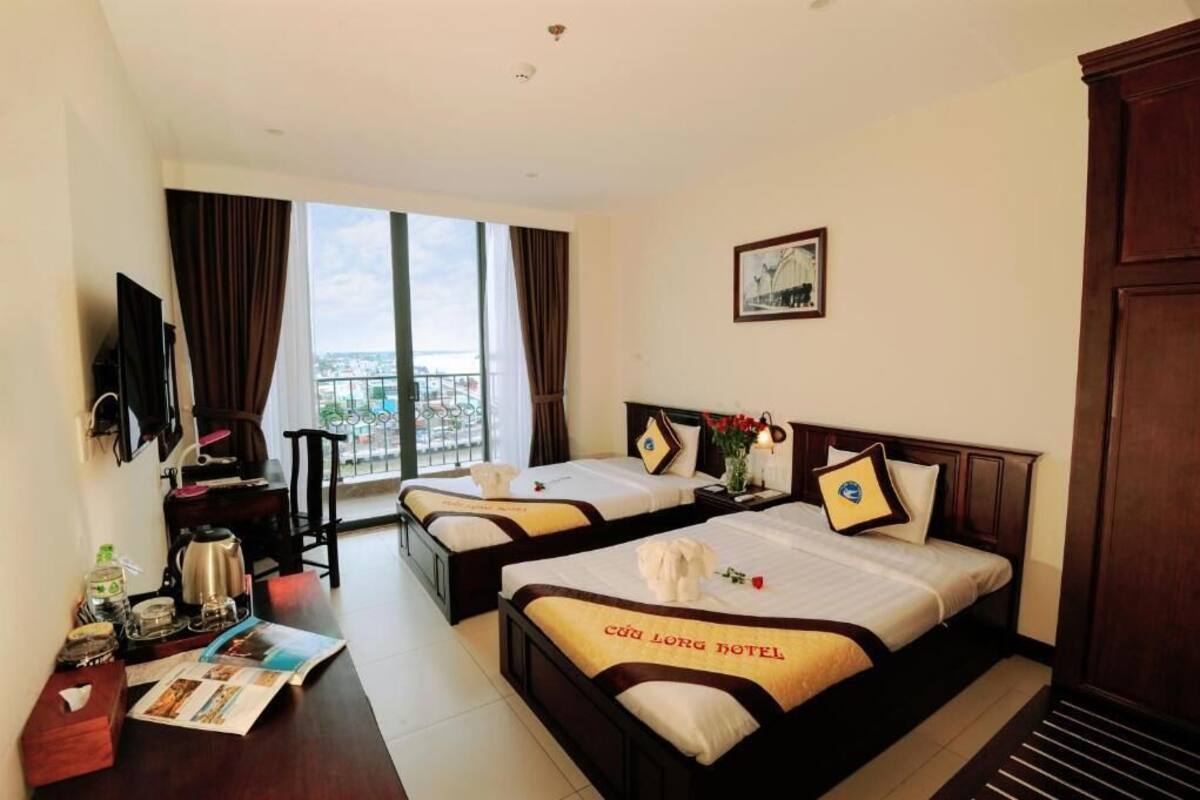 A hotel room with two beds, a TV, a desk with a chair, a window with curtains, and a balcony view.