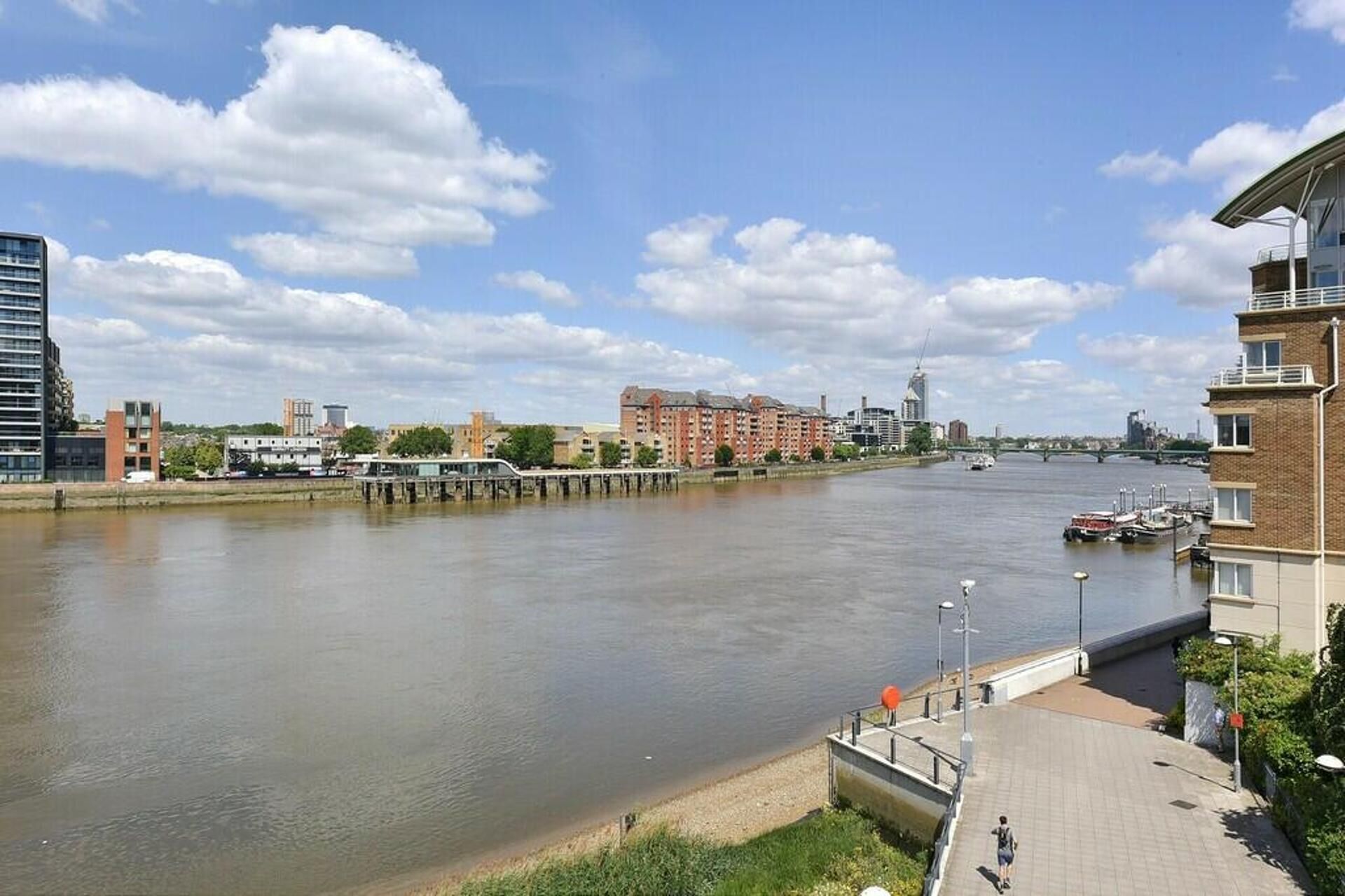 A riverside view with a pedestrian walkway, buildings, and a bridge in the background.