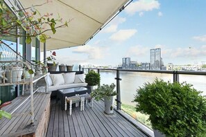 A deck with a sofa and table, overlooking a river with buildings in the background. - Luxury City Apartment in London Battersea Reach (London)
