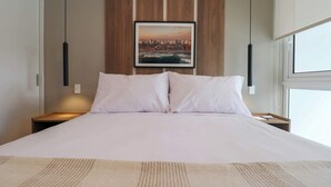 A neatly made bed with white linens, a framed cityscape picture on the wall, and a window on the right side.