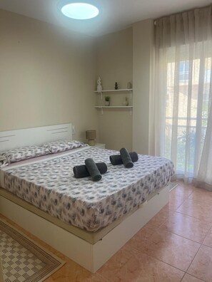 A bedroom with a bed, a bedside table, a lamp, a window with curtains, and a shelf with decorative items. - Rosaleda Monterey (Dénia)