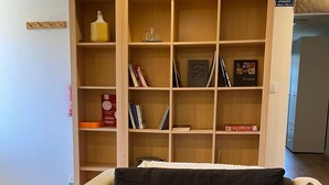 A living room with a beige sofa, two dark cushions, and a wooden bookshelf filled with books and decorative items.