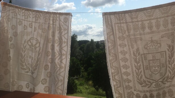 A view through a window with patterned curtains, a wooden ceiling, and a view of a green landscape with trees and a cloudy sky.