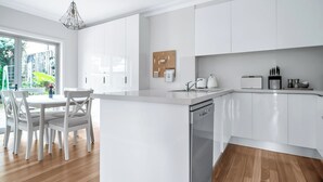 A modern kitchen with white cabinetry, a dishwasher, and a dining area with white chairs and a table. - The Newtowner - Stylish Home in the heart of Newtown! (Newtown)