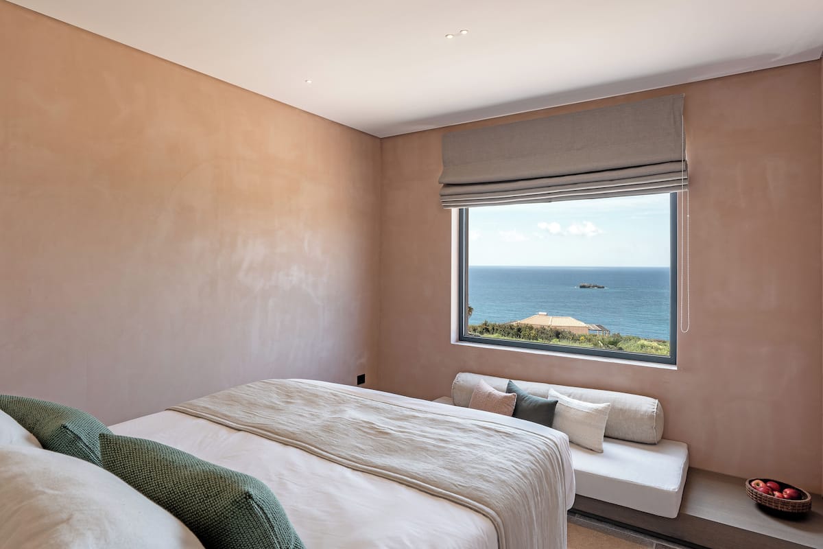 A bedroom with a large window, a bed, a bench, and a bowl of fruit.