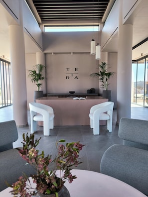 Un área de recepción moderna con un diseño minimalista, que incluye un mostrador de recepción, dos sillas blancas, plantas en macetas y un amplio ventanal con vistas.