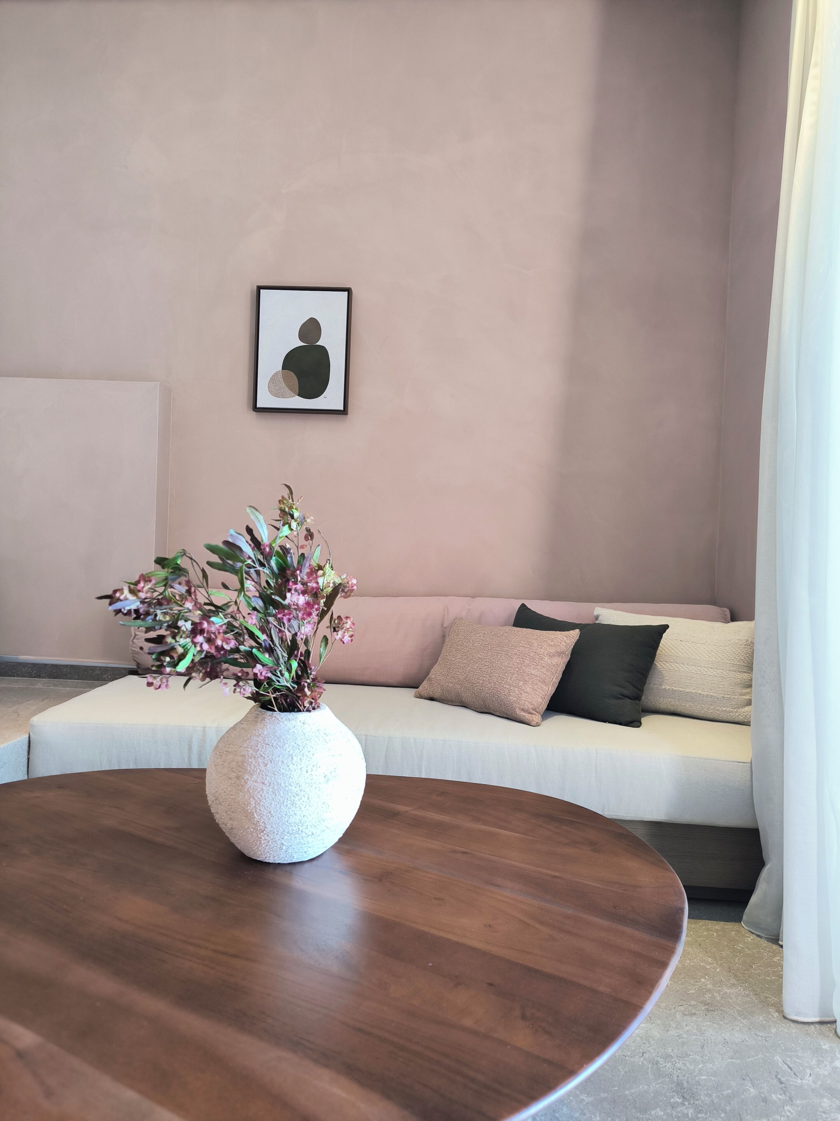 A living room with a round wooden table, a white sofa with cushions, a framed abstract artwork on the wall, and a vase with flowers on the table.