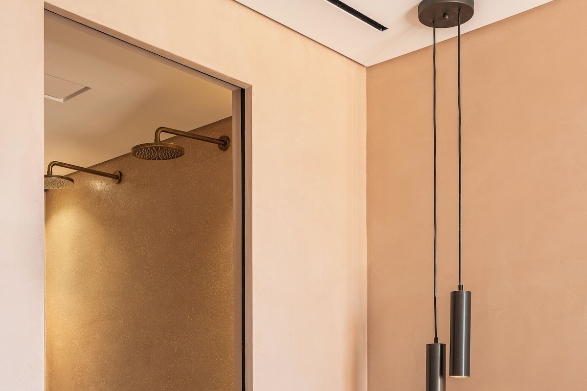 A modern hotel room with a glass shower enclosure, a bed with white linens, a bedside table with a vase, and two pendant lights.