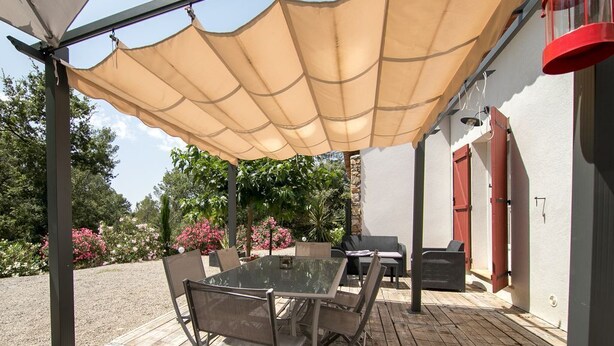 Patio esterno con tavolo e sedie riparato da un tetto, circondato da un giardino con fiori e alberi.