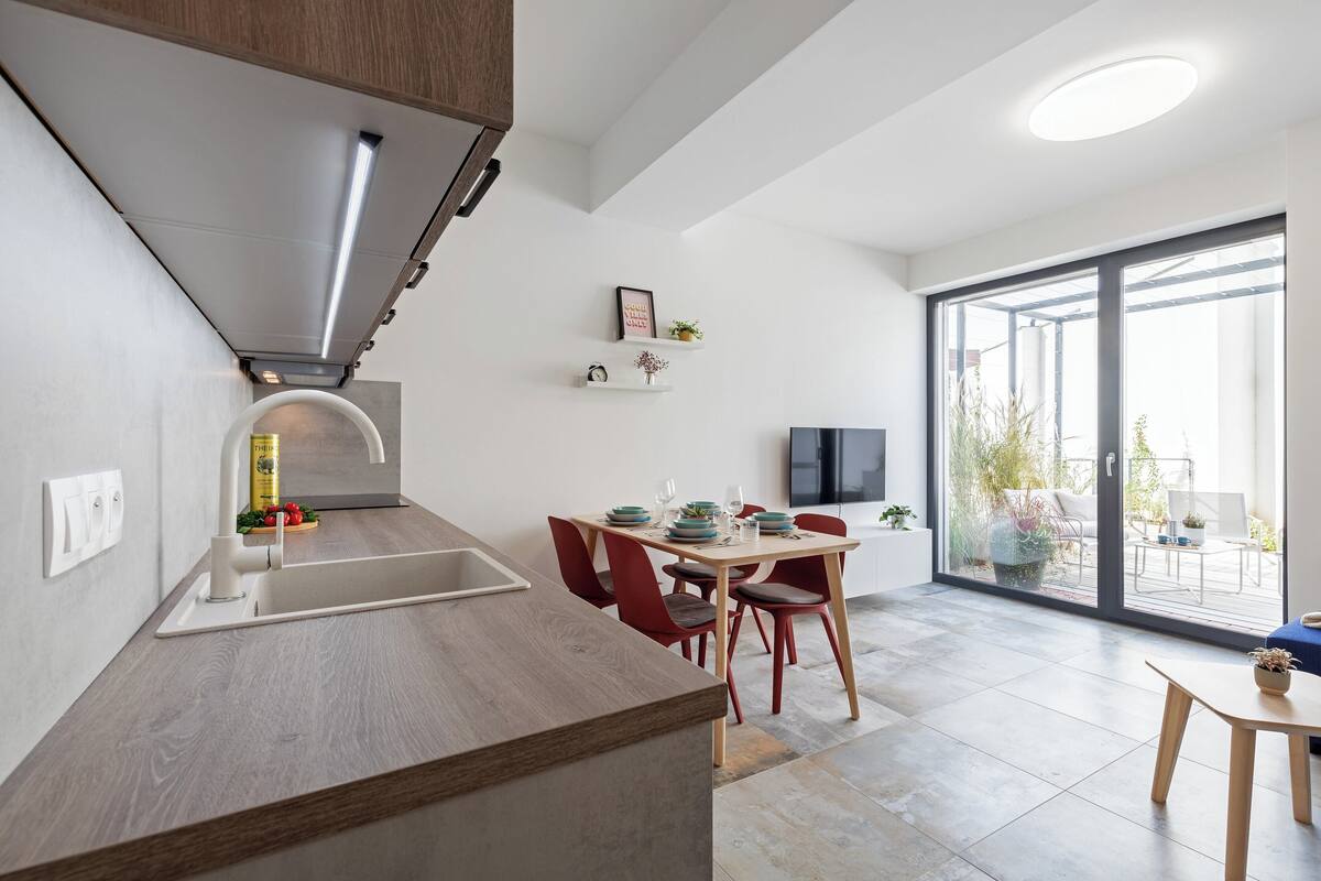 A modern kitchen with a built-in stove, a dining area with red chairs, and a large sliding glass door leading to a balcony.