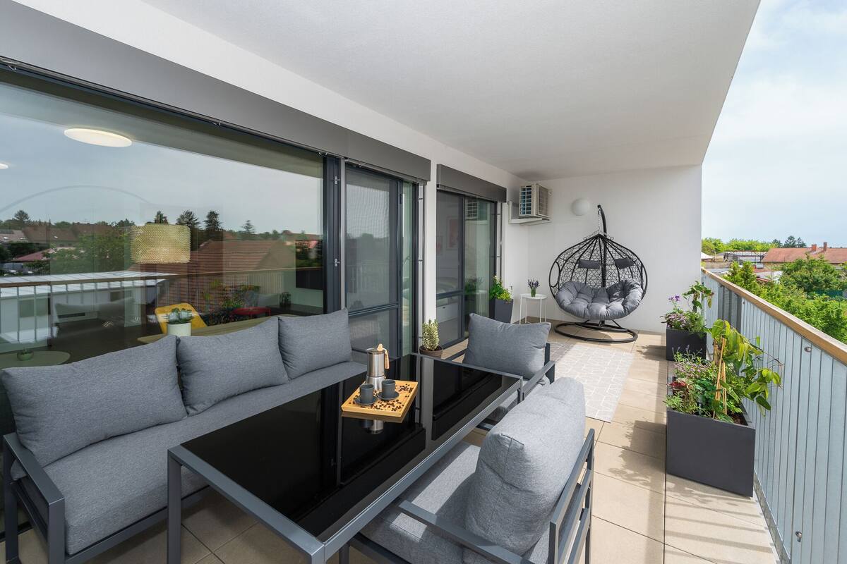 A balcony with a sofa, a table, and a hanging chair. There are potted plants and a view of the city.