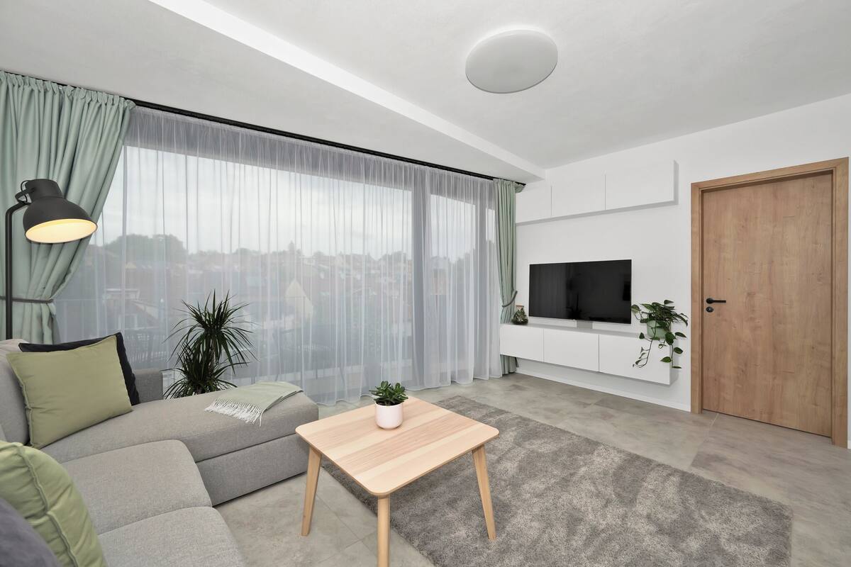 A modern living room with a sofa, a small wooden table, a TV on a white cabinet, and a view of the city through sheer curtains.