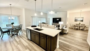 A modern kitchen with a central island, dining area with chairs, a sofa, and a TV in the living room. - 5 O`Clock Somewhere - 3/2 w/ Golf Cart in The Village of Lake Denham (The Villages)