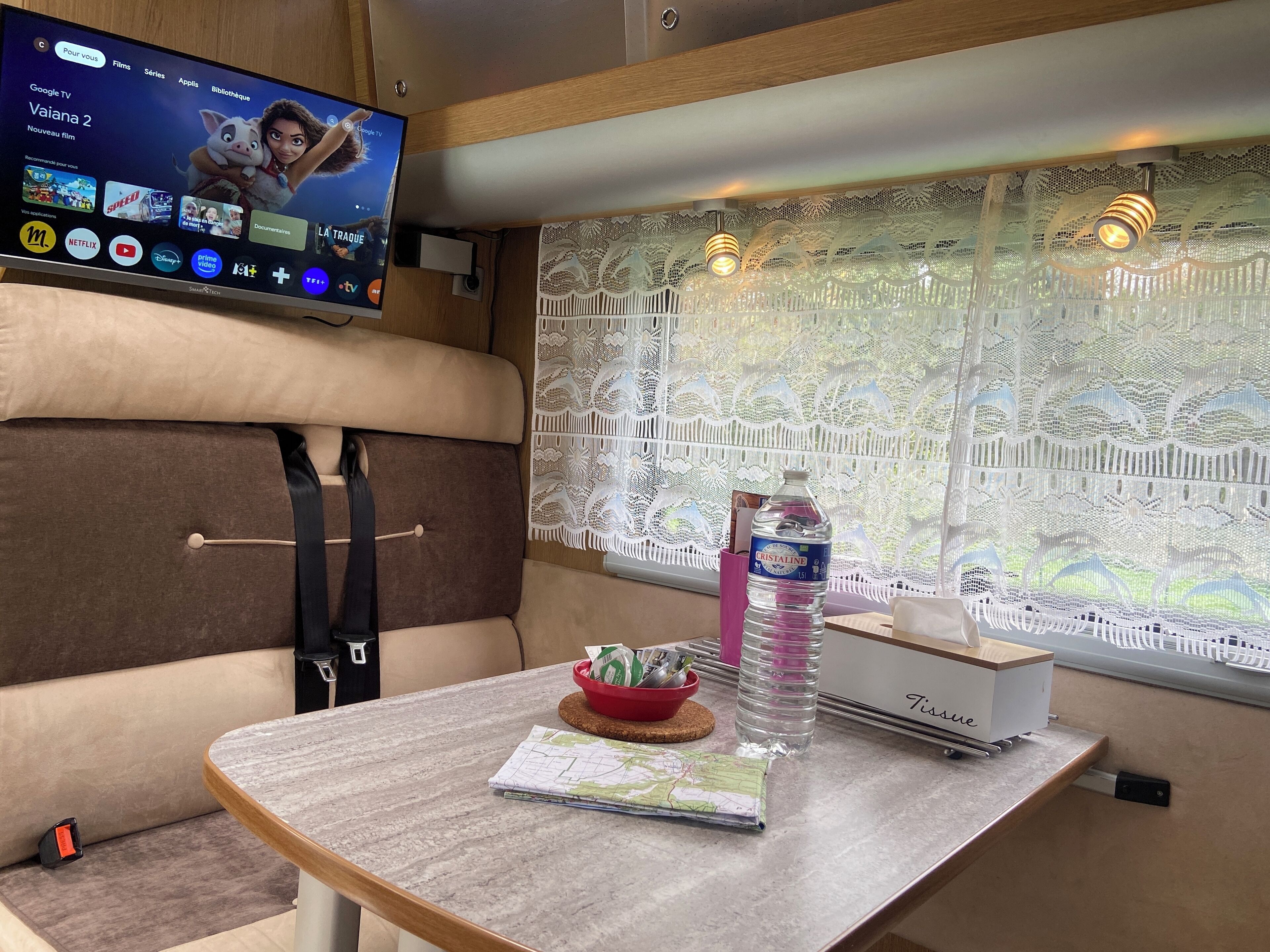 A cozy room with a TV displaying a streaming service, a table with a map and a water bottle, and a window with lace curtains.