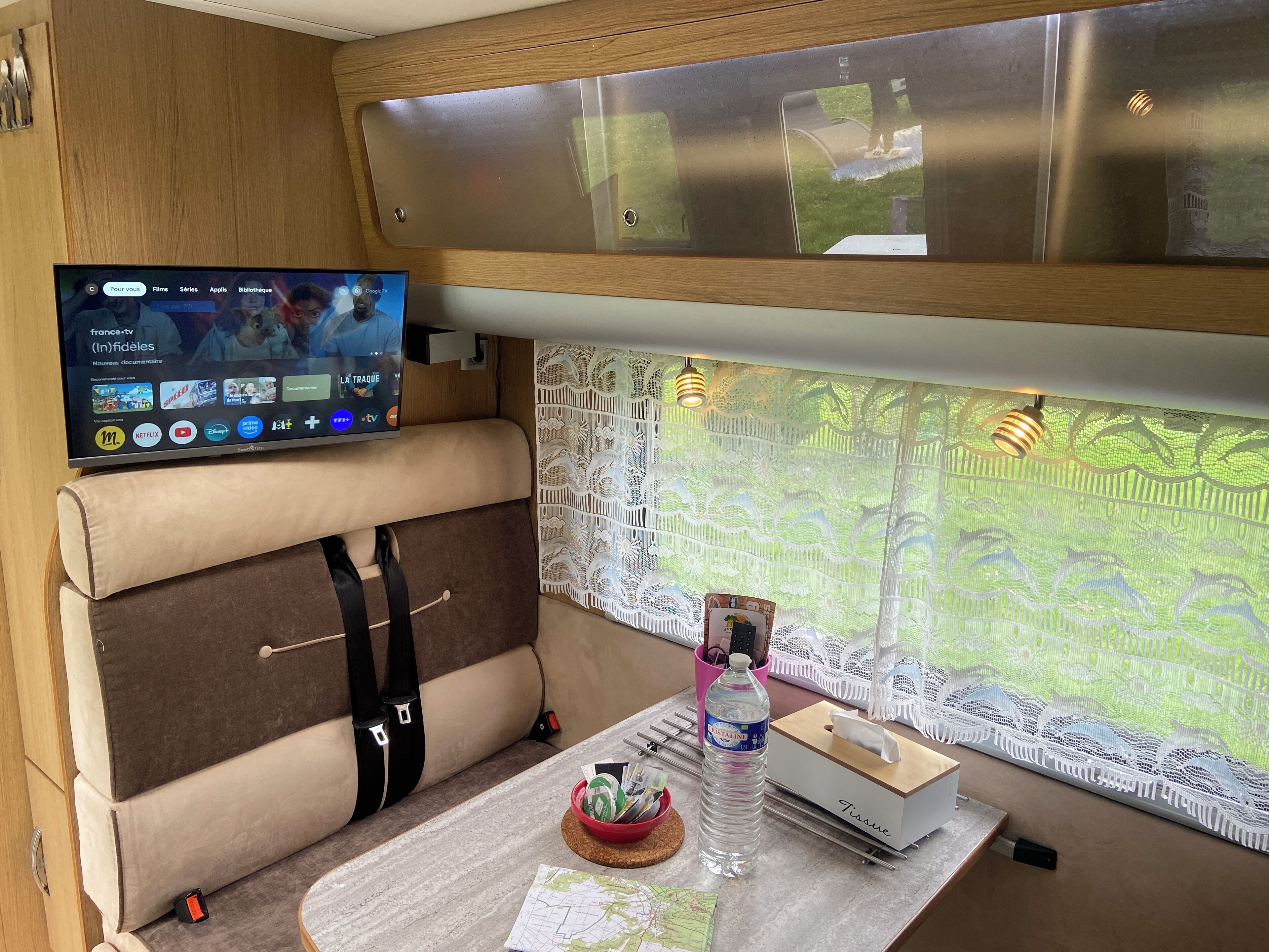 Ein gemütliches Interieur mit Fernseher, der einen Streaming-Dienst zeigt, einer gepolsterten Sitzecke, einem Fenster mit Spitzenvorhängen und einem Tisch mit Taschentuchbox und Flasche.